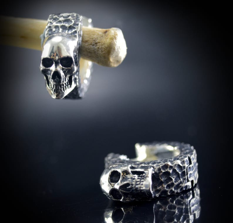 K&ouml;nnte beinhalten: Ein Paar silberne Totenkopf-Creolen mit strukturierter Oberfl&auml;che. Die Ohrringe werden auf einer Holzoberfl&auml;che gezeigt.