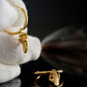 May include: Gold hoop earrings with a small bird skull charm hanging from each hoop.