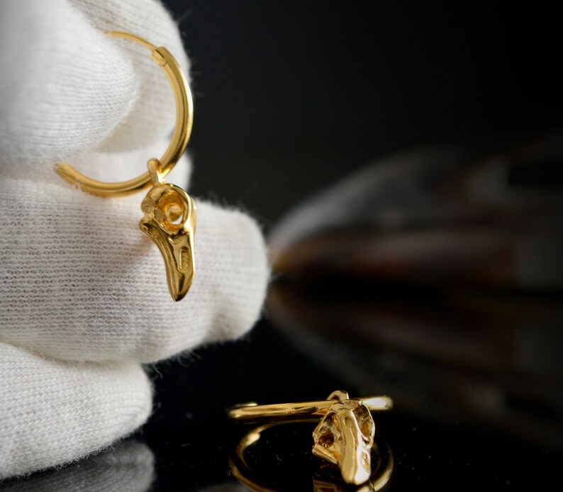 May include: Gold hoop earrings with a small bird skull charm hanging from each hoop.