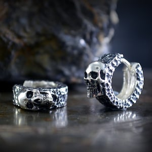 K&ouml;nnte beinhalten: Zwei silberne Totenkopf-Creolen mit strukturierter Oberfl&auml;che. Die Ohrringe sind aus Metall und haben einen Scharnierverschluss.