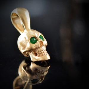 May include: A gold skull pendant with green gemstone eyes. The skull is detailed with a textured surface and a small loop at the top for attaching a chain.