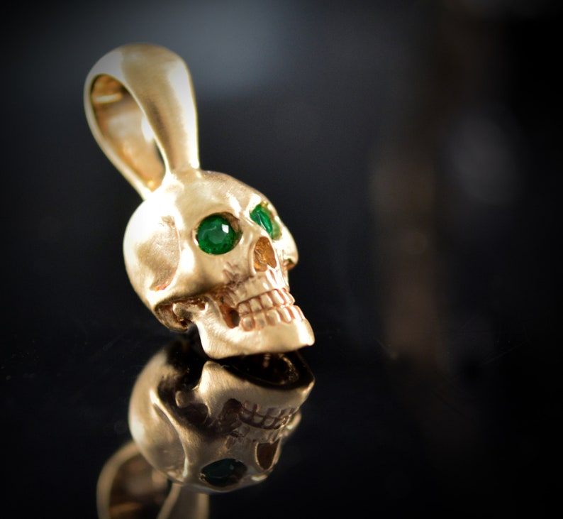 May include: A gold skull pendant with green gemstone eyes. The skull is detailed with a textured surface and a small loop at the top for attaching a chain.