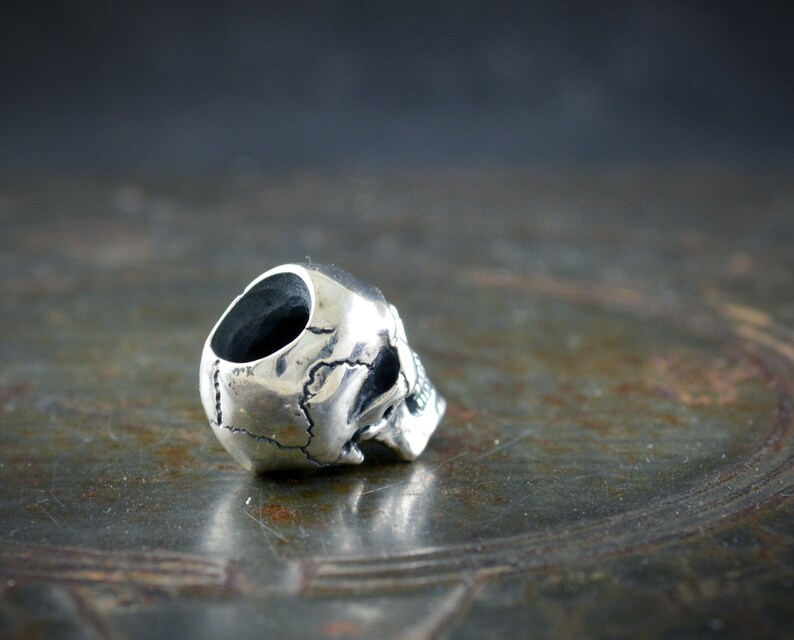 Op de afbeelding: Een zilveren schedelkraal met een gedetailleerd ontwerp. De kraal heeft een gat aan de bovenkant om op een armband of ketting te rijgen.