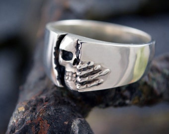 Op de afbeelding: Een zilveren ring met een schedelontwerp aan de ene kant en een handontwerp aan de andere kant. De ring ligt op een donker, getextureerd oppervlak.