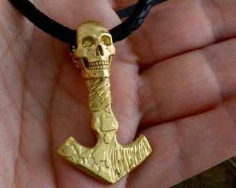 May include: A gold-toned pendant necklace featuring a skull with a detailed face and a textured hammer design. The necklace is on a black leather cord.