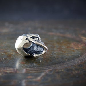Op de afbeelding: Een zilveren schedelkraal met een uitgeholde binnenkant, ontworpen voor gebruik op een armband of ketting.
