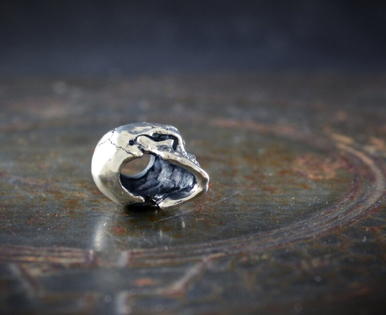 Op de afbeelding: Een zilveren schedelkraal met een uitgeholde binnenkant, ontworpen voor gebruik op een armband of ketting.