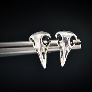 Può includere: Un paio di orecchini in argento a forma di teschio di uccello, ogni orecchino ha la forma di un teschio di uccello con un becco appuntito.