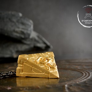 Peut inclure: Pendentif doré avec un motif d'œil stylisé, de forme carrée, suspendu à une chaîne sombre. Le design complexe du pendentif est mis en valeur sur un fond sombre et texturé, accentuant sa couleur dorée.