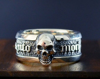 Op de afbeelding: Een zilveren ring met een schedelontwerp en de woorden "Memento Mori" gegraveerd op de band.