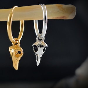 May include: Two hoop earrings with a skull charm. The earring on the left is gold and the earring on the right is silver.