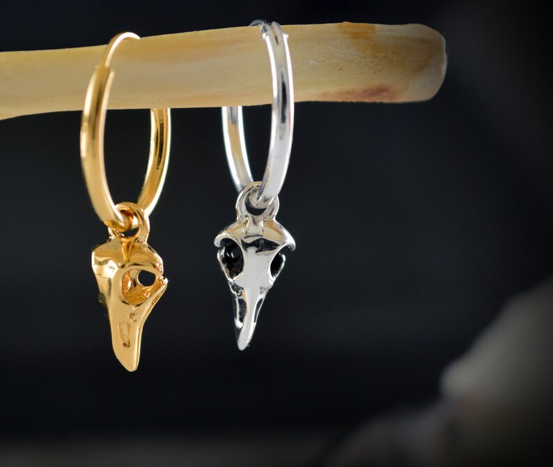 May include: Two hoop earrings with a skull charm. The earring on the left is gold and the earring on the right is silver.