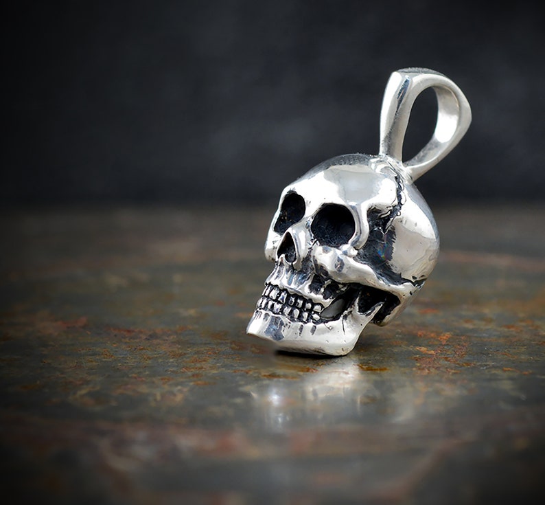 Pu&ograve; includere: Un ciondolo a forma di teschio in argento con un anello per una catena. Il teschio &egrave; dettagliato con orbite, un naso e denti.