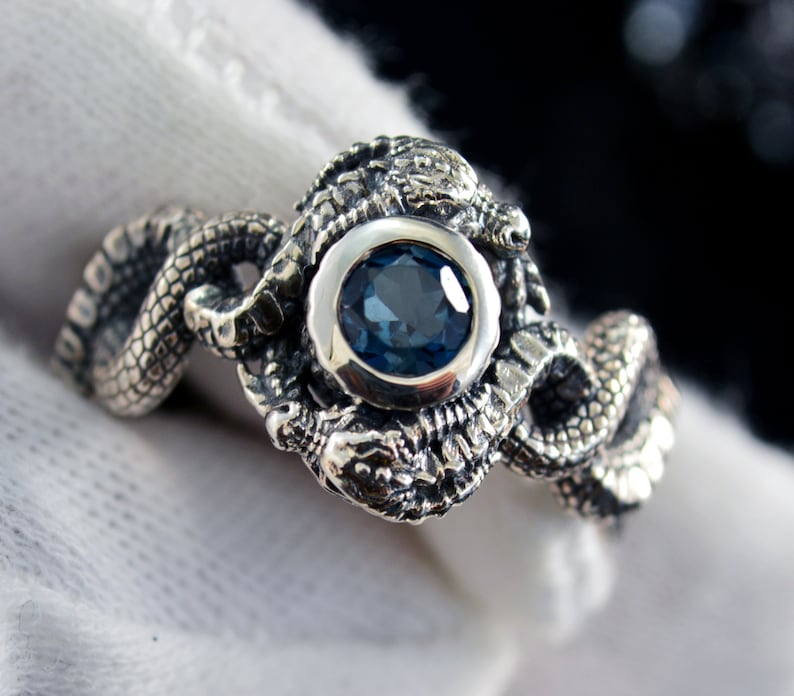May include: A silver ring featuring two intertwined snakes with a blue gemstone in the center.