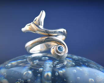 May include: A silver-toned ring with a spiral design, featuring a dolphin tail and a small, round, blue gemstone. The ring is set against a blurred, blue background, creating a sense of depth and focus on the jewelry.