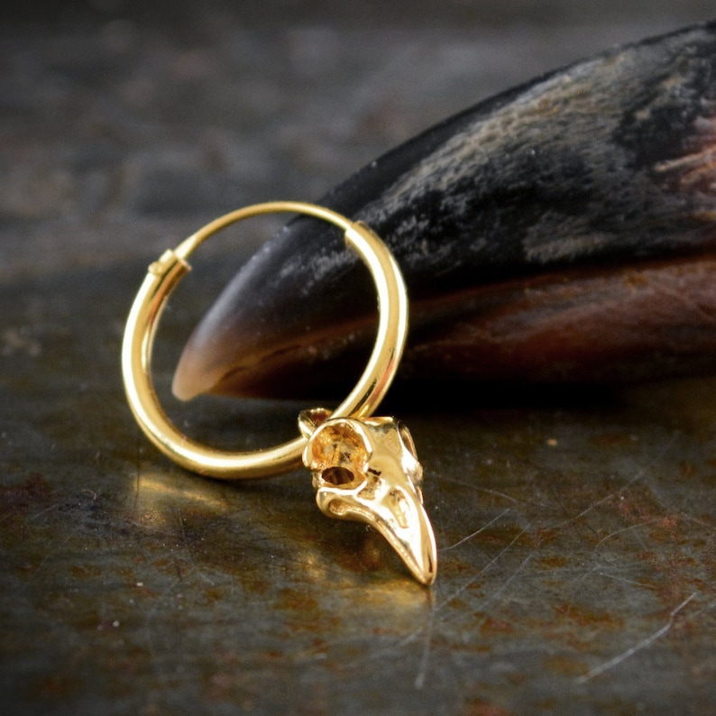 May include: Gold hoop earring with a small bird skull charm hanging from it. The earring is on a dark brown surface.