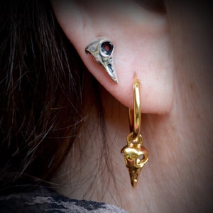May include: A pair of silver and gold earrings featuring a bird skull design. The silver earring is a stud, while the gold earring is a hoop with a dangling bird skull charm.