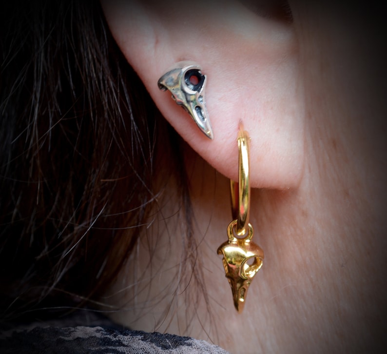 May include: A pair of silver and gold earrings featuring a bird skull design. The silver earring is a stud, while the gold earring is a hoop with a dangling bird skull charm.