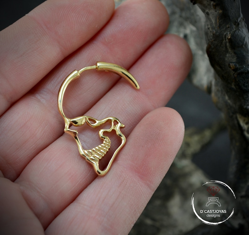 Op de afbeelding: Gouden schedelvormige oorring met een getextureerd ontwerp. De oorring is open en heeft een gebogen staaf die door het oor gaat.