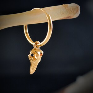 May include: Gold hoop earring with a small gold bird skull charm hanging from it.