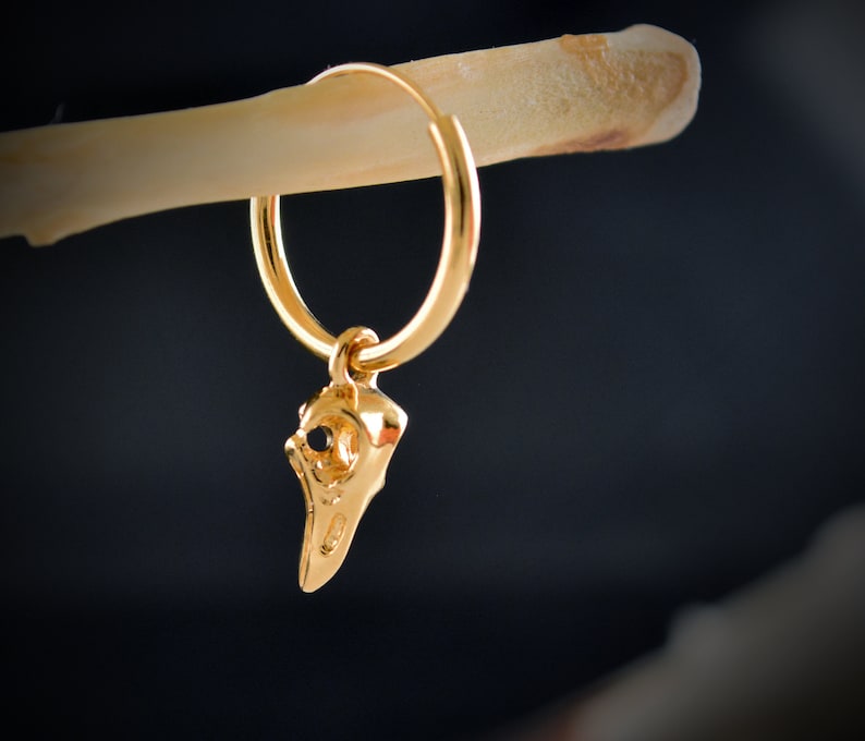May include: Gold hoop earring with a small gold bird skull charm hanging from it.