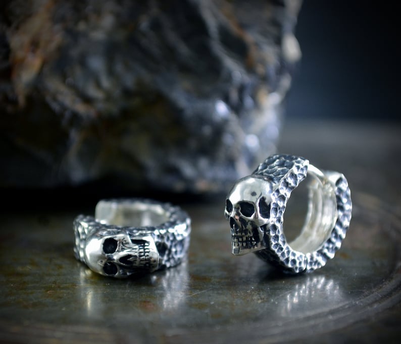 K&ouml;nnte beinhalten: Zwei silberne Totenkopf-Creolen mit strukturierter Oberfl&auml;che. Die Ohrringe sind aus Sterlingsilber gefertigt und haben ein Totenkopf-Design.