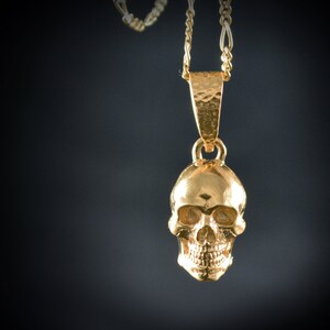 May include: A gold skull pendant necklace with a delicate chain. The skull is detailed with eye sockets, a nose, and a mouth.