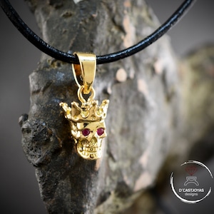 May include: Gold skull pendant necklace with a crown and red gemstone eyes. The necklace is on a black leather cord.