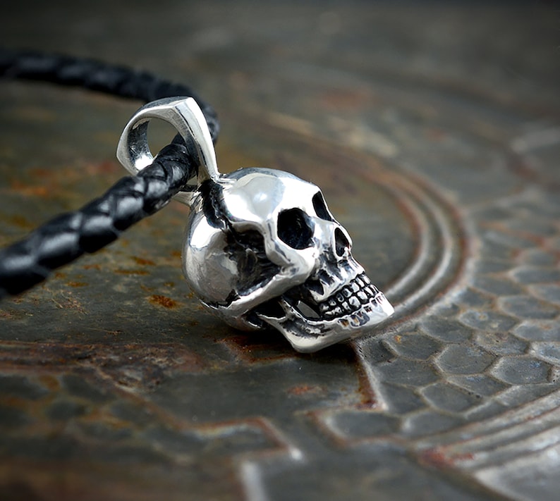Pu&ograve; includere: Un ciondolo a forma di teschio in argento con un design dettagliato, appeso a un cordino in pelle nera.