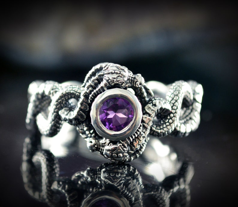 May include: A silver ring with a purple gemstone set in a silver bezel. The ring is designed with two intertwined snakes, each with a detailed head and scales.