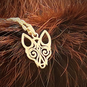 Peut inclure: Un collier avec un pendentif en forme de tête de loup orné de détails et de volutes complexes. Le pendentif est suspendu sur un fond de fourrure brune.