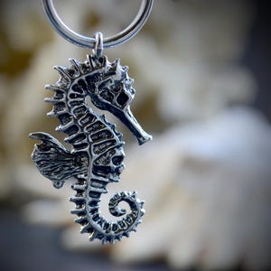 Puede incluir: Un colgante de plata detallado con forma de caballito de mar en un llavero. El caballito de mar tiene detalles intrincados, incluyendo un cuerpo texturizado, aletas puntiagudas y una cola enroscada. El llavero es un simple anillo plateado.
