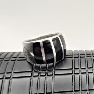 May include: A silver ring with a wide band featuring three vertical black rectangular inlays separated by thin silver lines. The ring has a polished finish and a bold, geometric design. The ring is a statement piece.