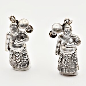 May include: A pair of silver-toned screw-back earrings featuring three-dimensional figures of sumo wrestlers. Each figure is depicted in a standing position, wearing a textured mawashi belt with a patterned design, and includes a small silver ball accent.