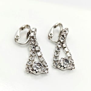 May include: A pair of silver-toned clip-on earrings. Each earring features a triangular design with a central, large, clear rhinestone and smaller rhinestones along the edges. The clip-on mechanism is visible at the top.