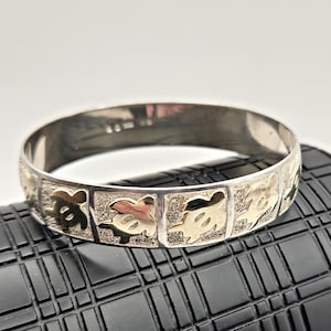Könnte beinhalten: Silbernes Armreif-Armband mit einem sich wiederholenden Muster aus goldfarbenen Meeresschildkröten-Designs. Das Armband hat eine polierte Oberfläche und wird auf einer strukturierten schwarzen Oberfläche präsentiert.