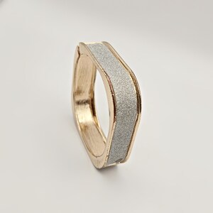 May include: A gold-toned bracelet with a rectangular shape and a silver glitter center. The bracelet has a smooth, polished finish on the gold sections. The design is modern and geometric, suitable for everyday wear or special occasions.