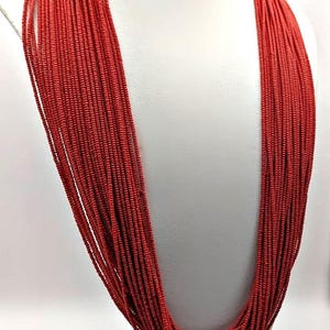 May include: A multi-strand necklace composed of numerous thin, red strands. The necklace is draped over a white mannequin bust, showcasing its cascading design. The strands are tightly packed, creating a full, textured appearance.