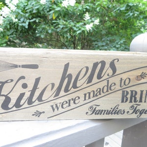May include: A wooden sign with the text "Kitchens were made to bring families together". The sign also features a whisk and a decorative flourish.