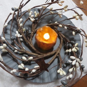 May include: A gray metal plate with a brown twig wreath decorated with white and gray berries. A small orange candle is in the center of the wreath.
