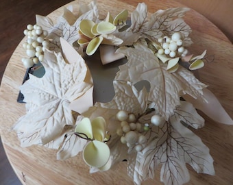 May include: A decorative wreath featuring cream-colored maple leaves, small white berries, and pale yellow flower accents. The wreath is arranged on a round, light brown wooden surface, creating a natural, autumnal theme.