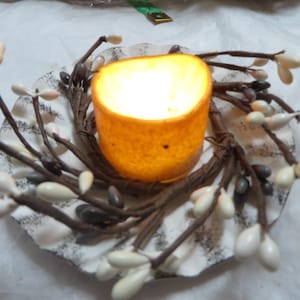 May include: A white ceramic candle holder with a rustic, distressed finish. The holder is decorated with a wreath of brown twigs and white berries. A small, round, orange candle sits in the center of the holder.