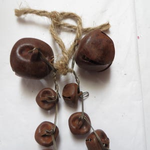 May include: A rustic, brown bell garland with a natural jute rope hanger. The garland features several small, round bells strung together with wire. The bells have a weathered appearance, suggesting an antique or vintage style.
