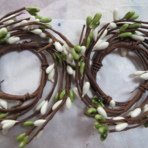 May include: Two decorative candle rings made of brown, twisted stems adorned with white and green berry-like accents. The rings are arranged on a white surface, creating a natural, rustic aesthetic. Ideal for seasonal home decor.