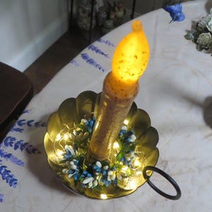 May include: A battery-operated yellow LED taper candle with a textured, speckled surface sits inside a gold-toned metal scalloped candle holder. The base is decorated with a small wreath of blue and white floral accents and warm fairy lights.