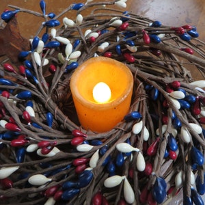 May include: A wreath made of brown twigs decorated with white, red, and blue berry-shaped ornaments. A lit candle sits in the center of the wreath.