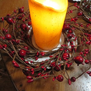 May include: A brown twig wreath with red berries surrounding a lit candle. The candle is a pillar candle with a speckled, off-white finish.