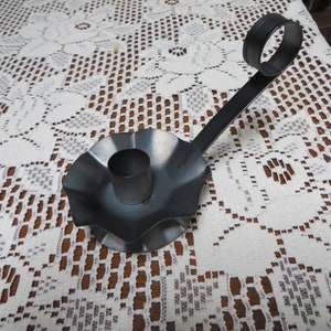 May include: A black metal candle holder with a fluted base and a curved arm. The holder is designed to hold a candle in the center of the base.