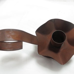 May include: Rustic metal candle holder with a wavy, flower-shaped base and a long, curved handle. The holder is a rusty brown color.
