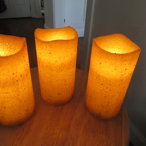 May include: Three pillar candles with a speckled, beige finish. The candles are lit and glowing.